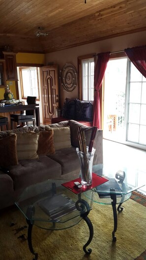 A living room with a glass coffee table, a sofa with cushions, a dining area with chairs, and a kitchen visible in the background.