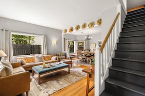 A living room with a sofa, ottoman, and coffee table, a dining area with chairs and a table, and a staircase with a wooden handrail.