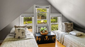 Un dormitorio con techo inclinado, ventanas amplias, cama con ropa de cama estampada, cómoda y planta en el alféizar.