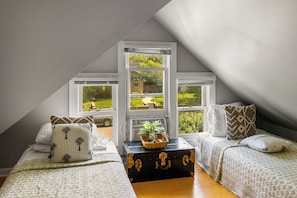 A bedroom with a sloped ceiling, large windows, a bed with patterned bedding, a chest of drawers, and a plant on the windowsill.