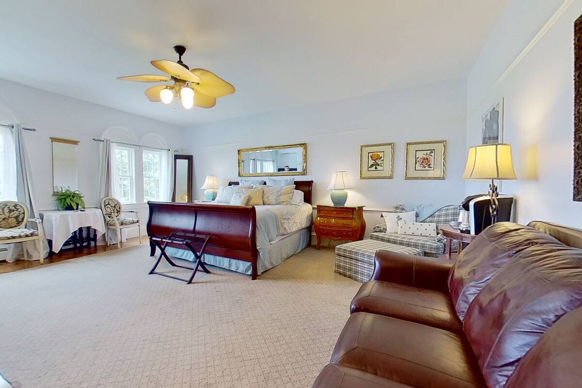 Une chambre spacieuse avec un grand lit, un coin salon avec un canapé en cuir marron, un bureau avec une chaise et un ventilateur de plafond.