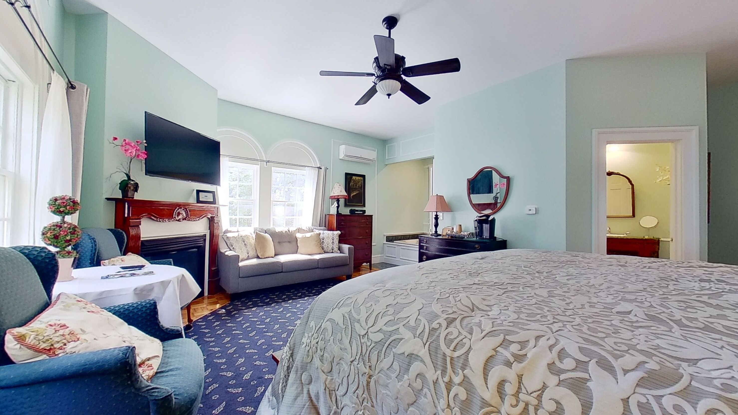 Une chambre d’hôtel avec un grand lit, un canapé, une télévision, un ventilateur de plafond et une salle de bain visible à travers une porte ouverte.
