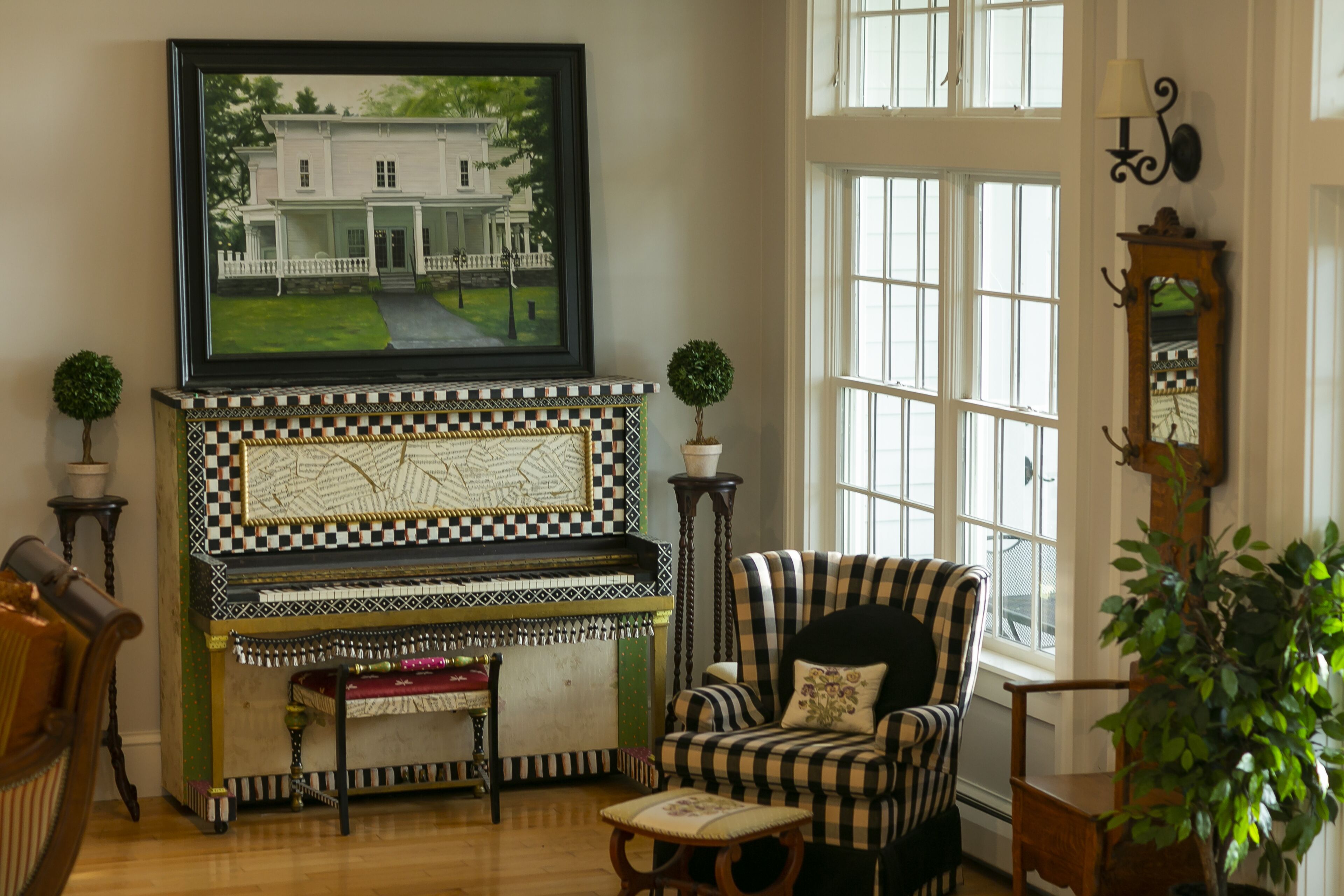 A room with a checkered fireplace, a framed painting of a house, a striped armchair, and a large window.