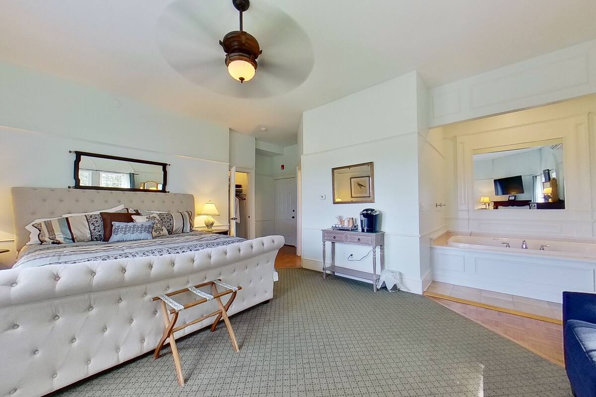 Une chambre spacieuse avec un grand lit, une table de chevet, une commode et une salle de bain équipée d’une baignoire et d’un grand miroir.