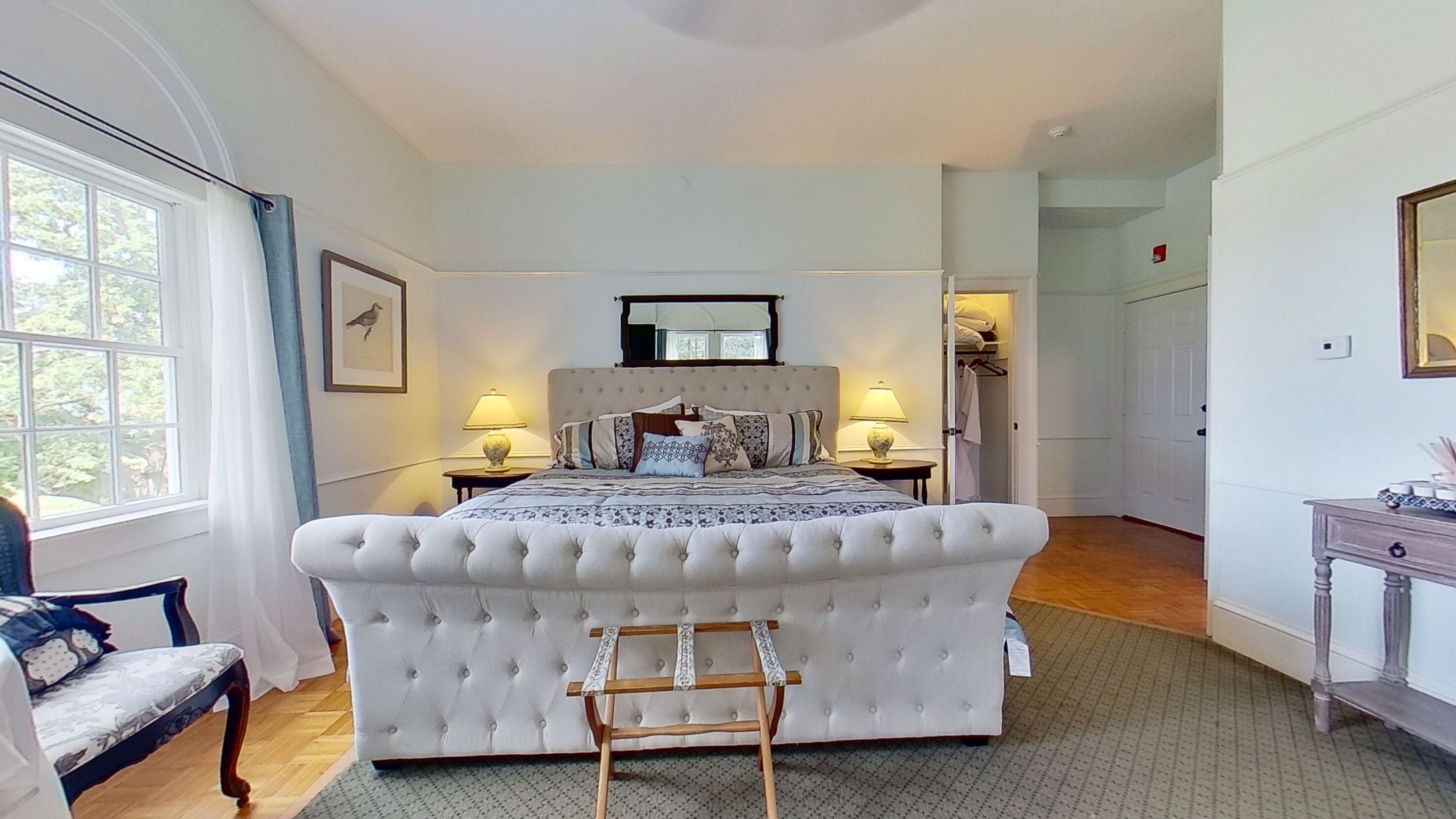 A spacious bedroom with a large bed, two bedside tables with lamps, a wooden chair, and a dresser.