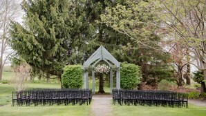 A wedding ceremony setup with a wooden arch and black chairs arranged in rows on a grassy path. - John Joseph Inn (Groton)