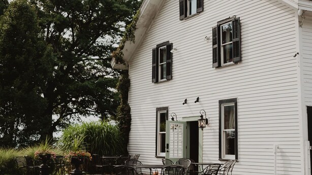 A white building with a gabled roof, multiple windows with shutters, and a stone wall in front. - John Joseph Inn (Groton)