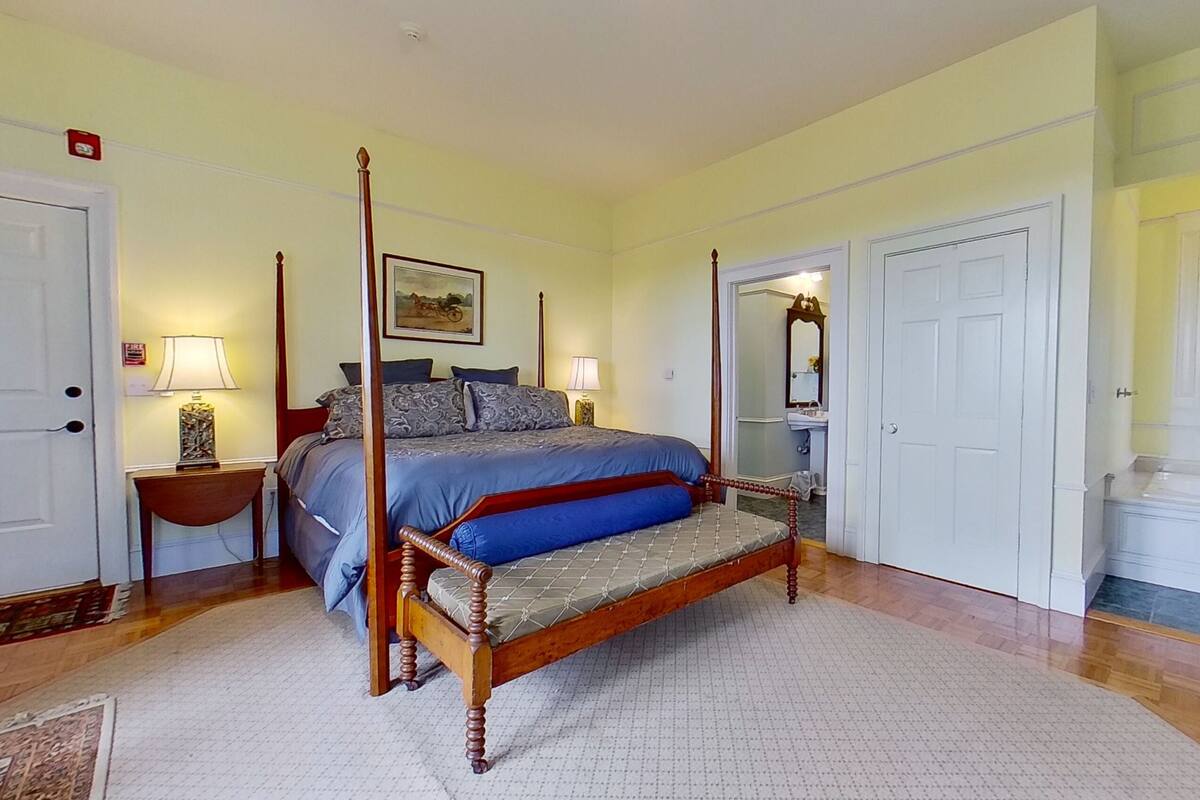 Un lit à baldaquin avec un cadre en bois et une couvre-lit bleue, une table de chevet avec une lampe, un tableau au mur et une salle de bain avec un miroir et un lavabo.