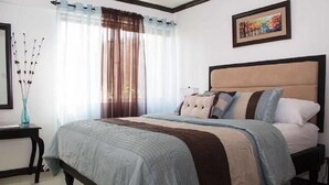 A neatly made bed with a headboard, a bedside table with a vase, and a window with sheer curtains. - Coconut Suite at Turtle Towers (Ocho Rios)
