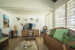 Una sala de estar con un sofá de mimbre, una mesa de centro de vidrio, un televisor y un adornos en la pared.