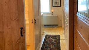 Un couloir intérieur en bois mène à une salle de bain dotée d’un lavabo blanc et d’un tableau encadré accroché au mur.