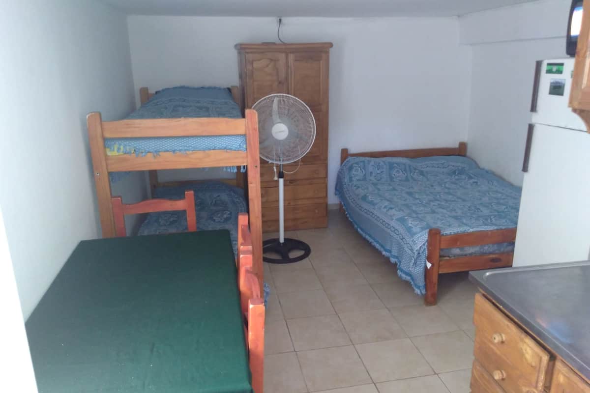A small, tidy room with two bunk beds, a single bed, a wooden wardrobe, a fan, and a small table.