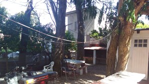 Un vehículo estacionado en un espacio al aire libre con sombra, con una mesa y sillas, rodeado de árboles y un edificio.