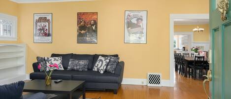 A living room with a black sofa, a coffee table, and a rug. There are posters on the wall and a dining area in the background.