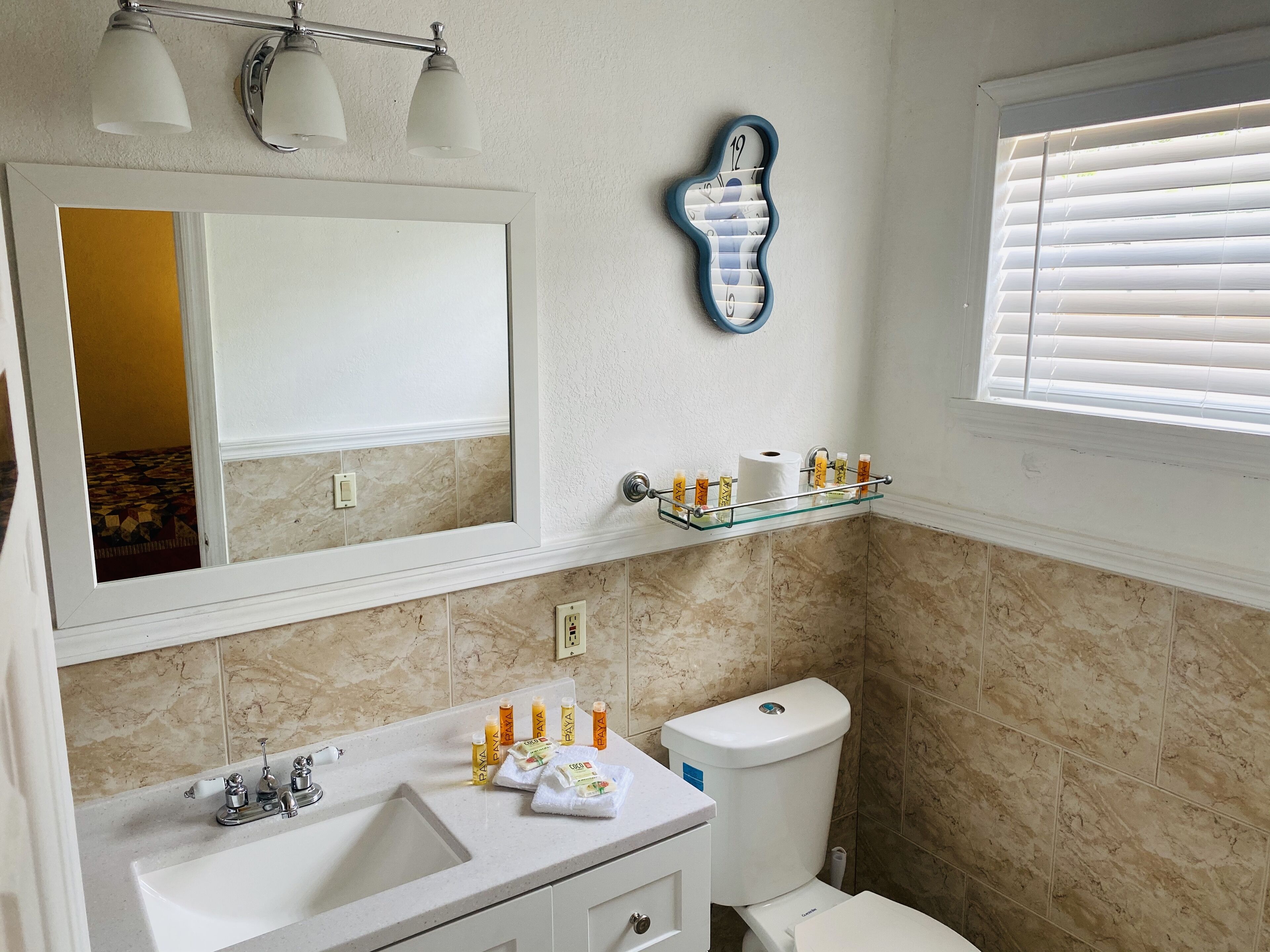 Une salle de bain avec un grand miroir, une étagère en verre où sont rangés les articles de toilette, des toilettes et une fenêtre avec des stores.
