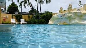 Piscina cristalina con una escultura de leones en pedestal, rodeada de follaje y una valla de madera.
