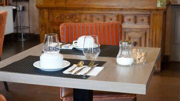 A dining table set with a white teapot, cups, and a sugar bowl, with a striped orange chair and a wooden cabinet in the background.