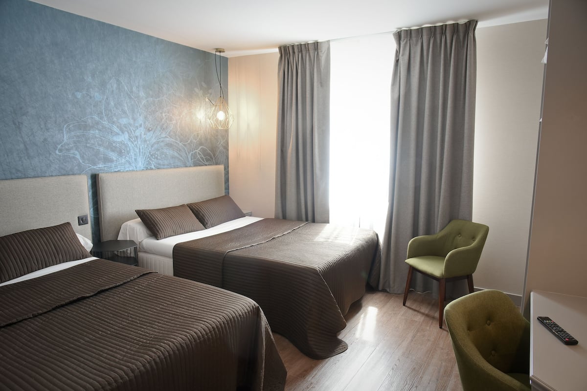 Une chambre d’hôtel avec deux lits, un fauteuil vert, une télécommande et une grande fenêtre avec des rideaux.