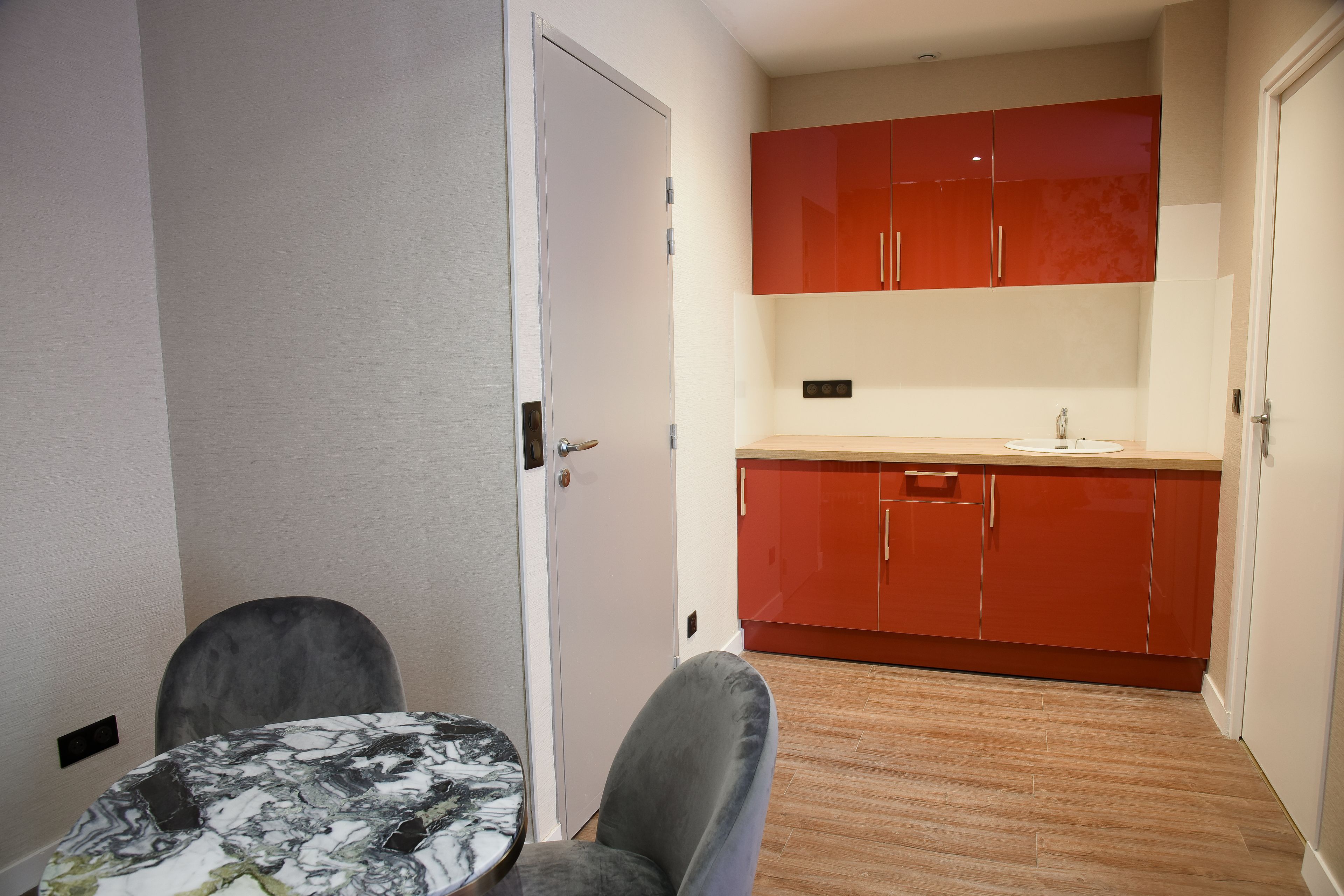 A compact kitchen with red cabinets, a marble-patterned table, and a grey chair.