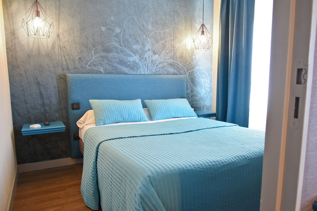 Un lit bien fait, avec une tête de lit bleue, une table de chevet sur laquelle se trouve une lampe, et un mur orné d’un motif floral.
