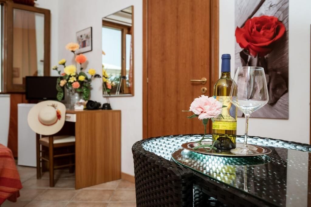 A room with a dining table set with a bottle of wine and a glass, a vase with flowers, a hat on a chair, and a wooden cabinet.