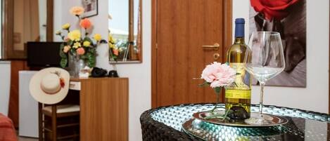 A room with a dining table set with a bottle of wine and a glass, a vase with flowers, a hat on a chair, and a wooden cabinet.