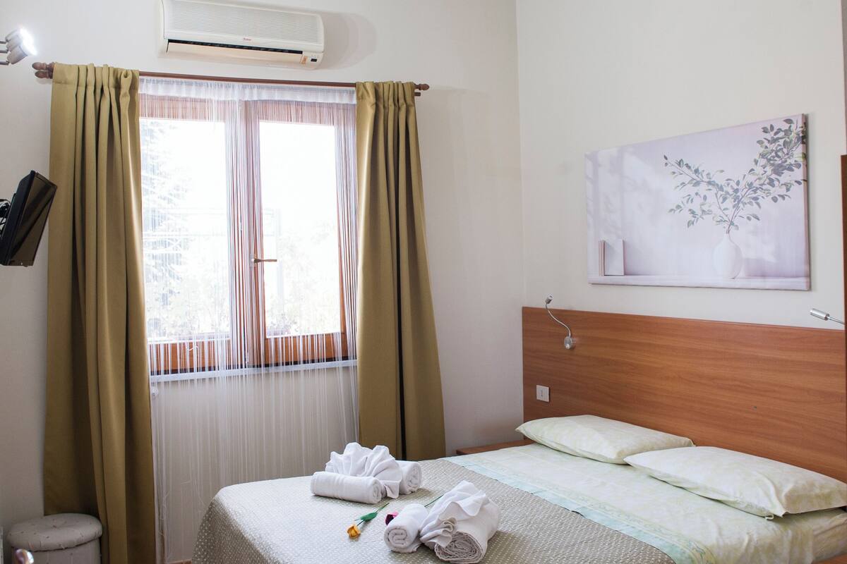 Habitación de hotel con cama, mesita de noche, silla, pequeña mesa, equipo de aire acondicionado, ventana con cortinas y una obra de arte colgada en la pared.