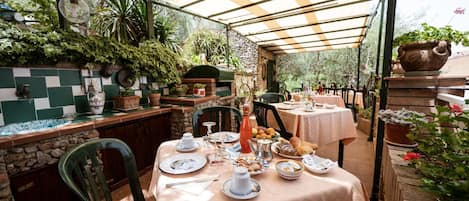An outdoor dining area with tables set for breakfast, surrounded by potted plants and a stone wall.