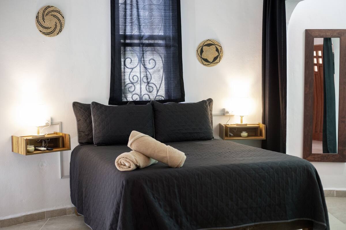 Un lit bien fait, avec une couvre-lit sombre et deux oreillers, une table de chevet avec une lampe et des œuvres d’art décoratives accrochées au mur.