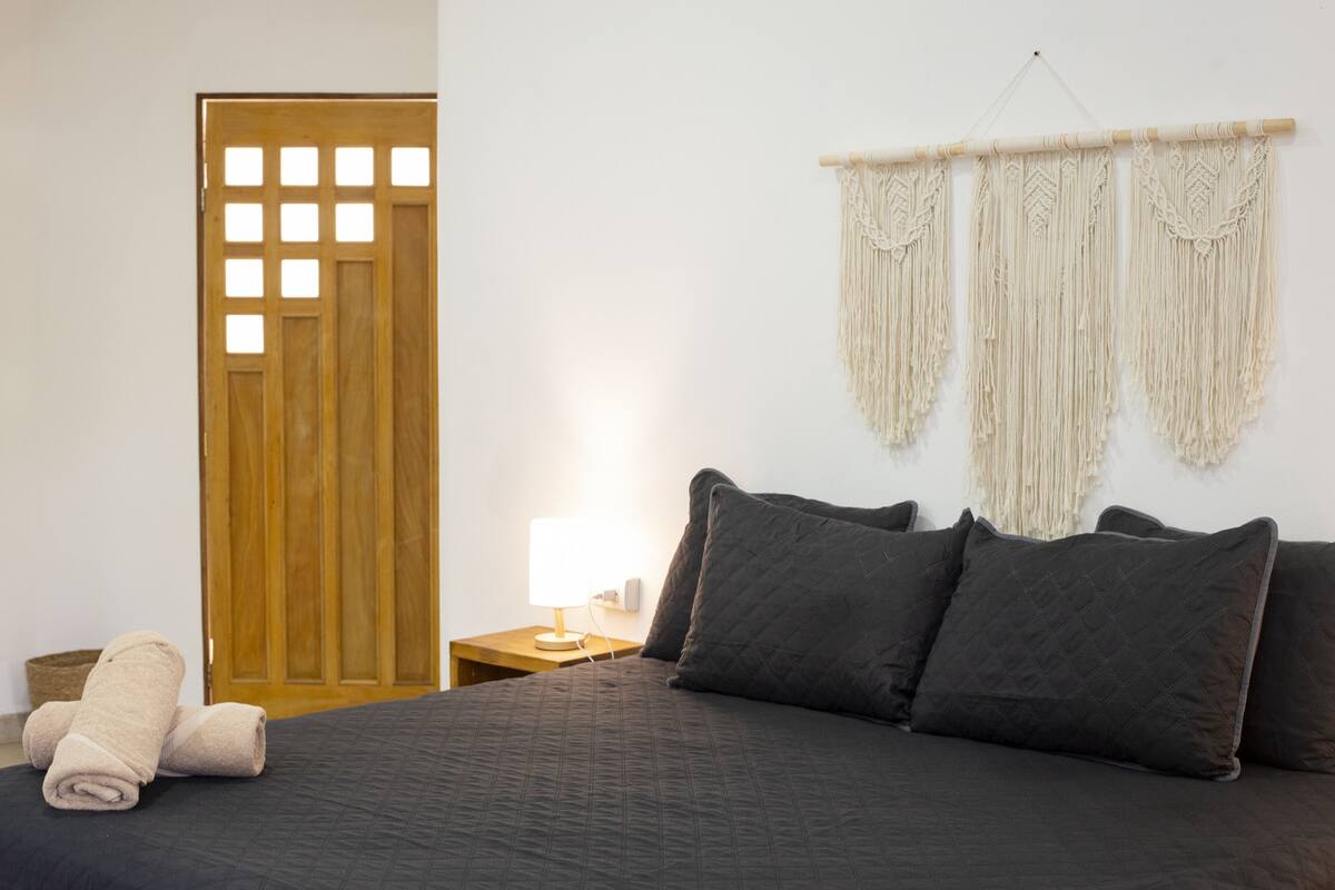 Un lit bien fait, avec une literie aux couleurs sombres, une porte en bois ornée d’un motif quadrillé et une décoration en macramé accrochée au mur.