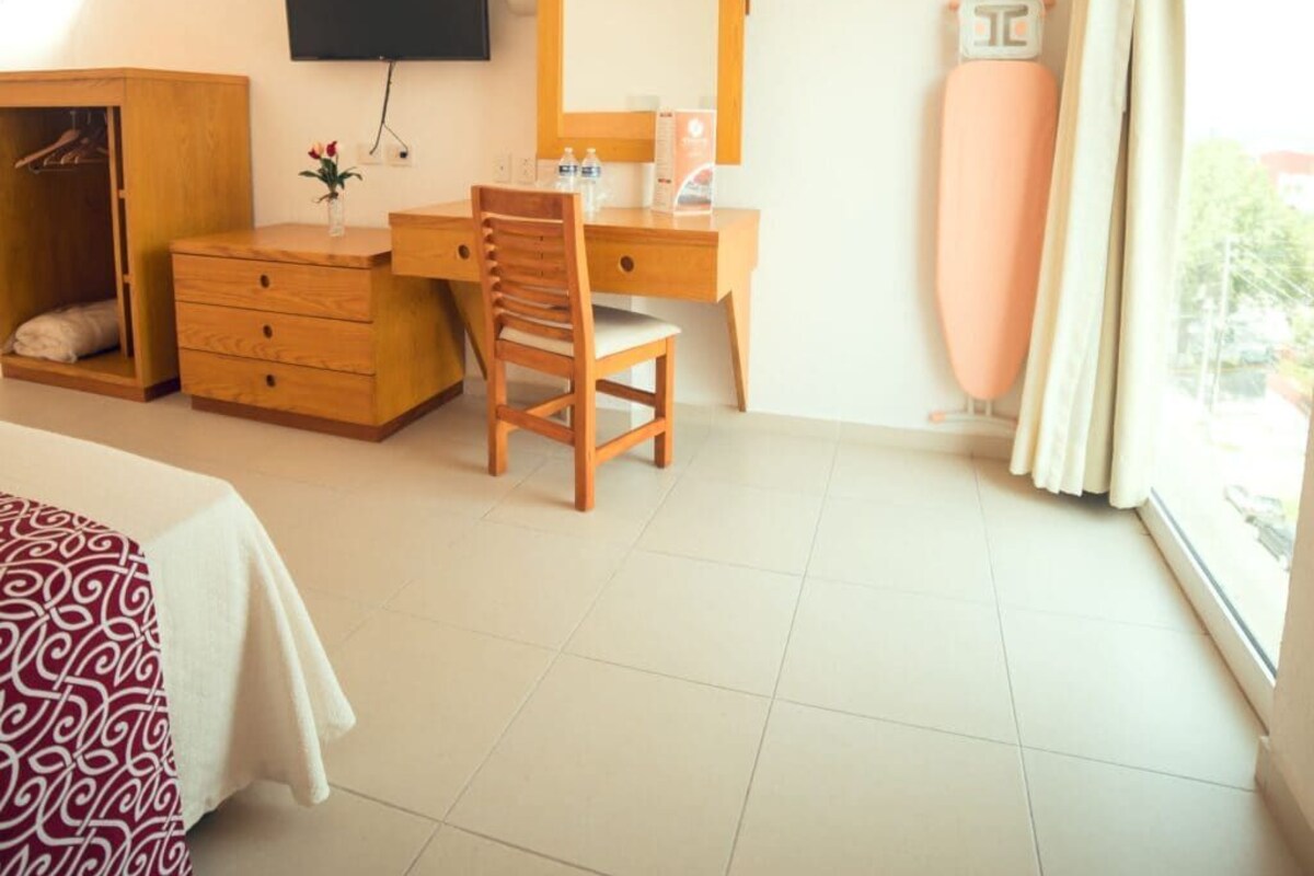 A hotel room with a bed, a wooden desk with a chair, a television mounted on the wall, a mirror, a radiator, and a window with curtains.