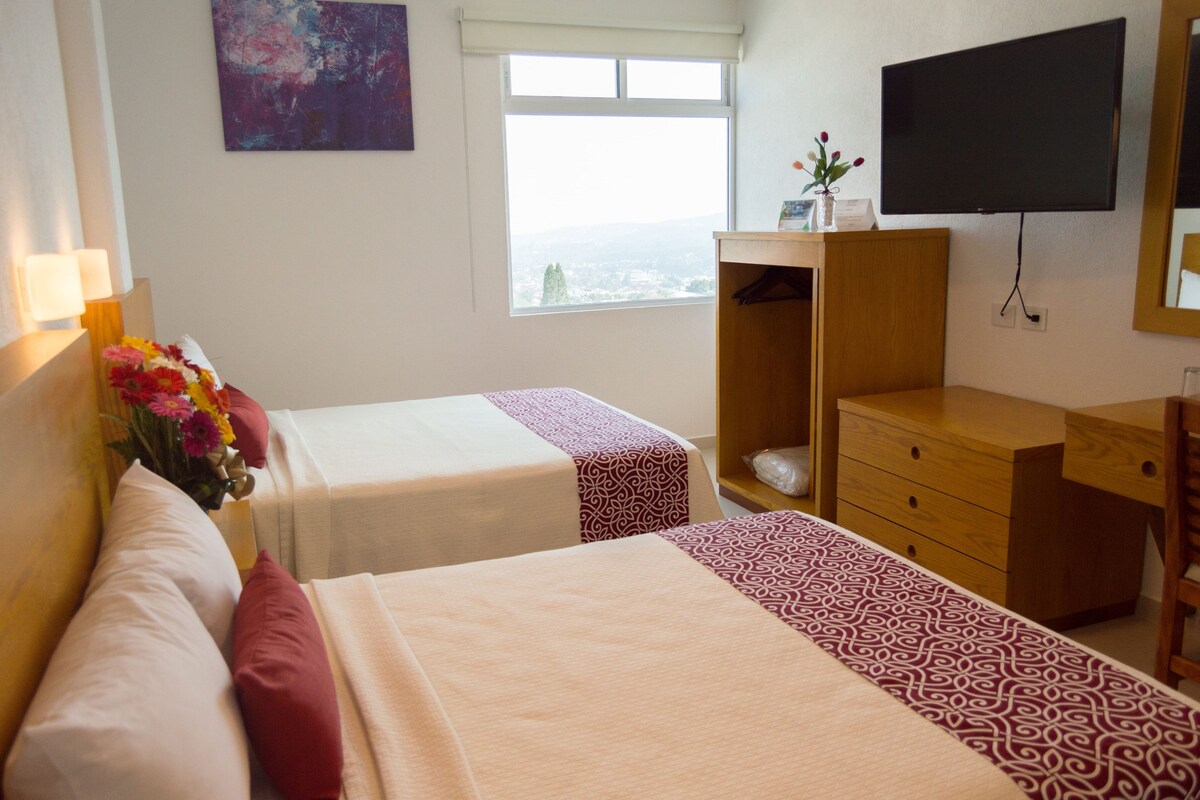 A hotel room with two beds, a dresser, a TV, a window with a view, and a vase of flowers.