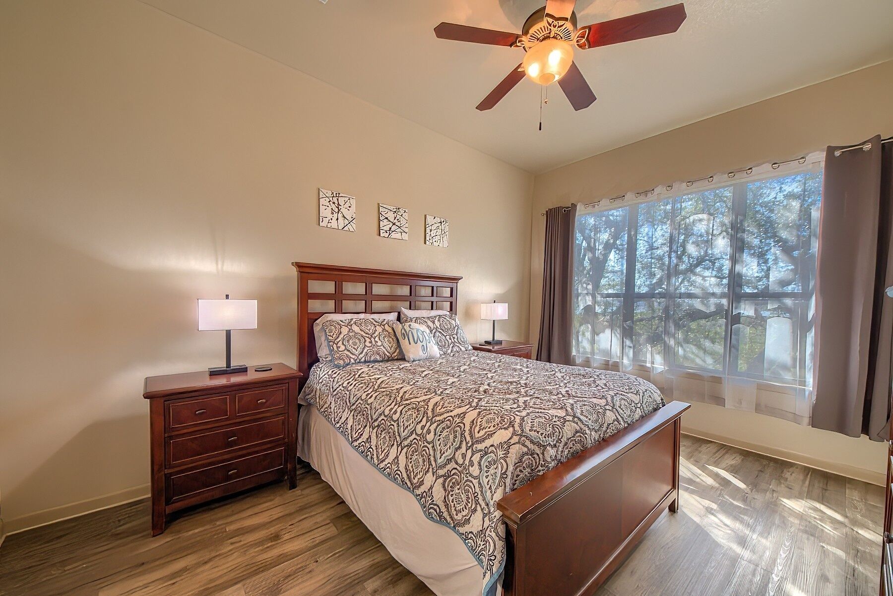 A bedroom with a wooden bed, nightstands, a ceiling fan, and a large window with curtains.