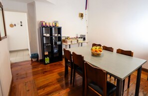 A dining area with a wooden table and chairs, a fruit basket, a black shelving unit with various items, and a small kitchen area with a coffee maker. - Guest House do Largo (Faro)