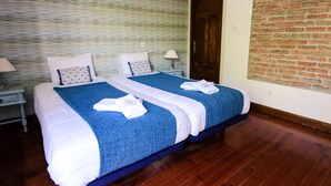 A double bed with a blue and white bedspread, two pillows, and two towels on the bed. There is a nightstand with a lamp and a small drawer. The room has a wooden floor and a brick wall. - Guest House do Largo (Faro)
