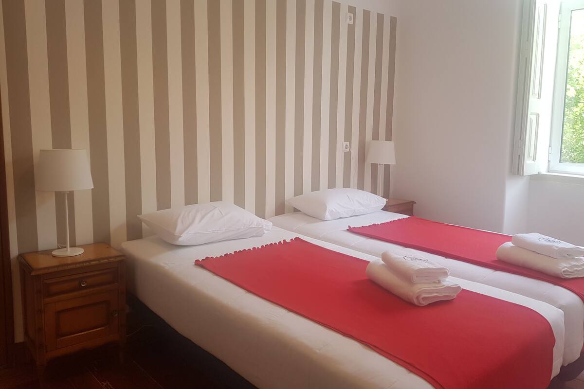 A hotel room with two beds, striped wallpaper, a wooden nightstand, and a window with a view.