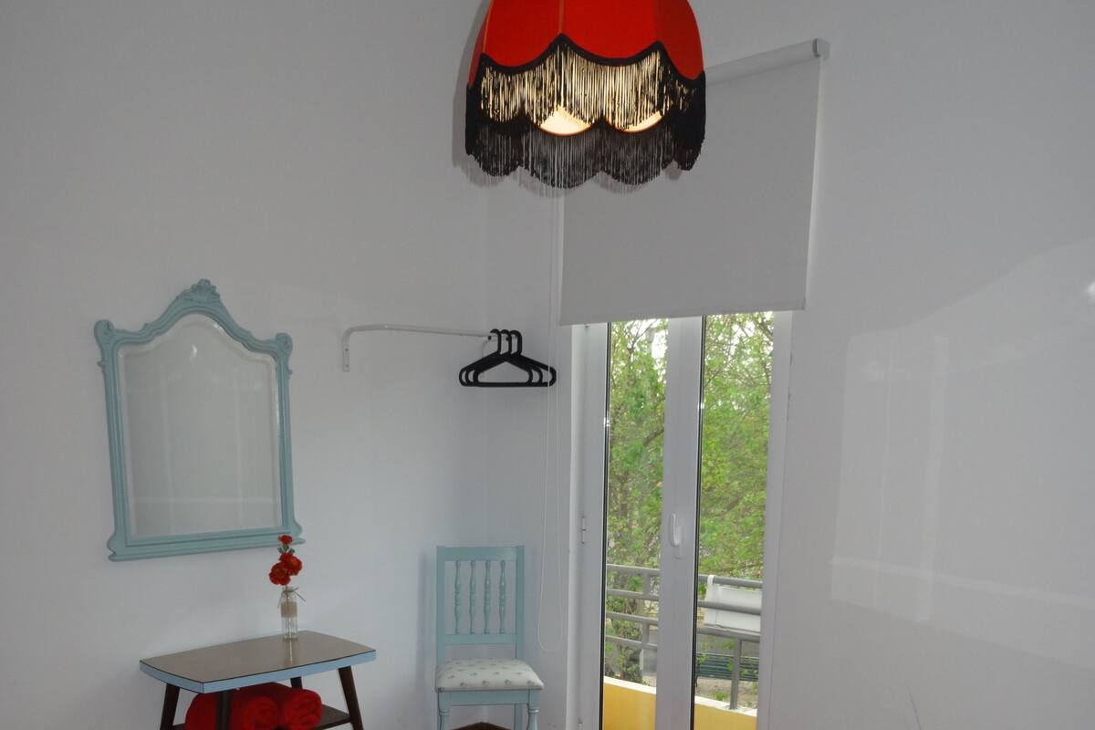A bedroom with a large bed, a small table with a mirror, a chair, and a hanging light fixture.