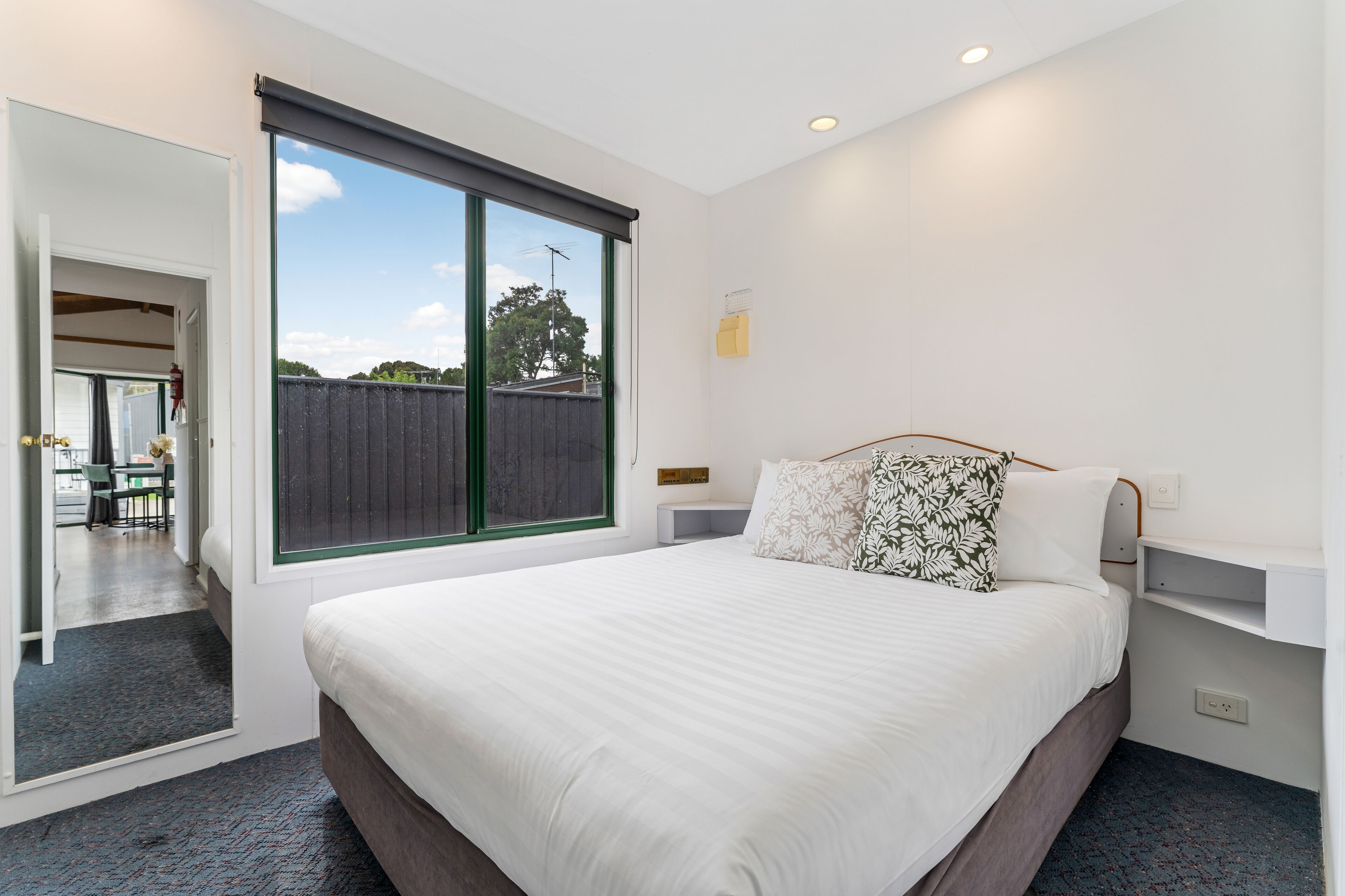 A bedroom with a large window, a bed with white bedding, a bedside table, and a view of the outside.