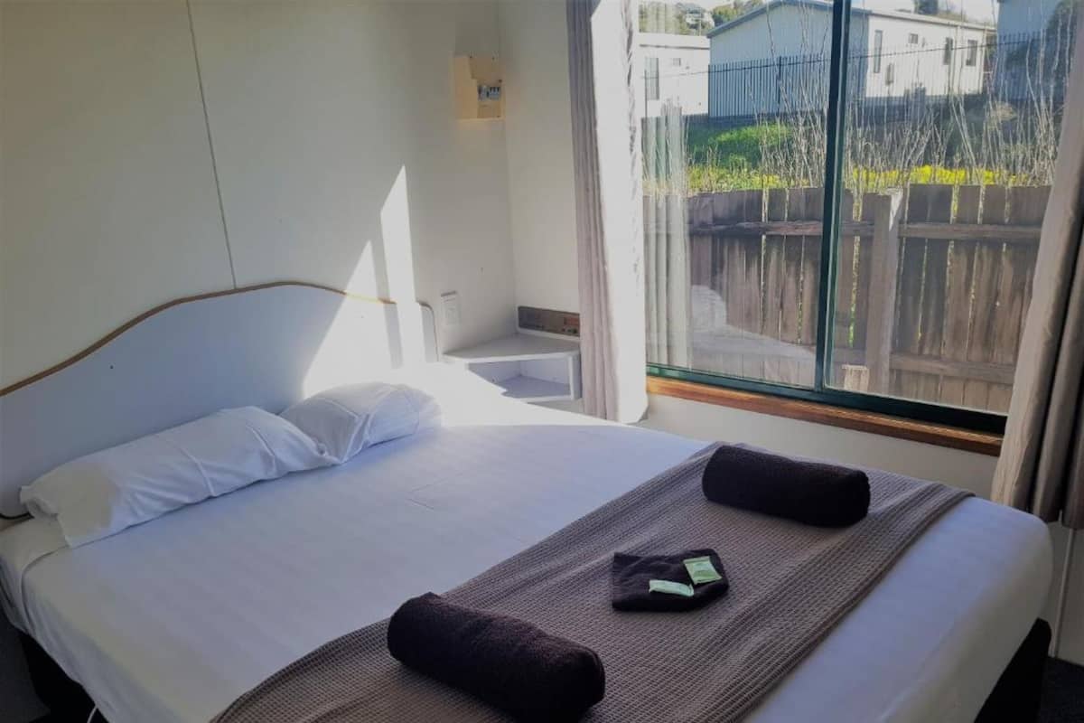 A hotel room with a bed, a small bedside table, a window with a view of the outside, and a wall-mounted light.