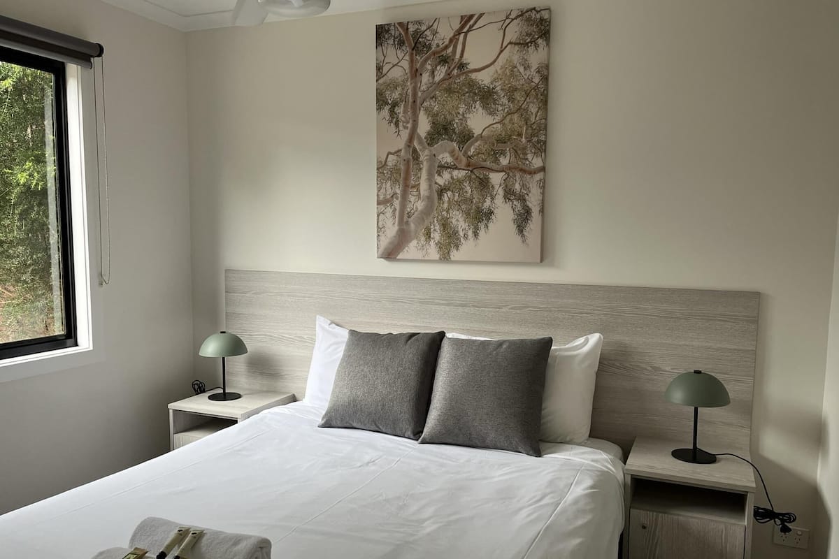 A neatly made bed with white linens, two bedside tables with lamps, and a large tree painting on the wall.