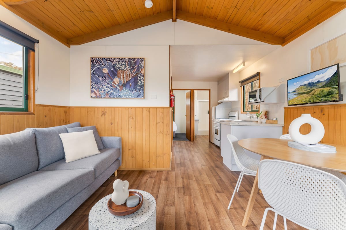 A cozy room with a wooden ceiling, a grey sofa, a dining table with chairs, and a kitchen area visible through an open door.