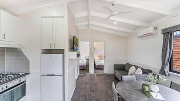 A compact kitchen with a refrigerator, stove, and cabinets. There is a dining area with a table and chairs, and a sofa with cushions. A ceiling fan and an air conditioning unit are visible.