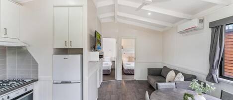 Cocina compacta con refrigerador, estufa y armarios. Hay un comedor con mesa y sillas, y un sofá con cojines. Se ve un ventilador de techo y un equipo de aire acondicionado.