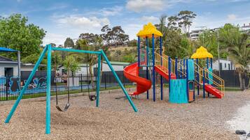 Un parc infantil amb cadiretes i tobogan, envoltat per una tanca i arbres.