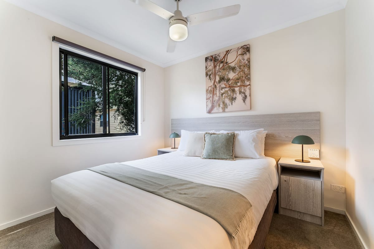 A bedroom with a bed, a nightstand, a lamp, a ceiling fan, a window, and a wall-mounted artwork.