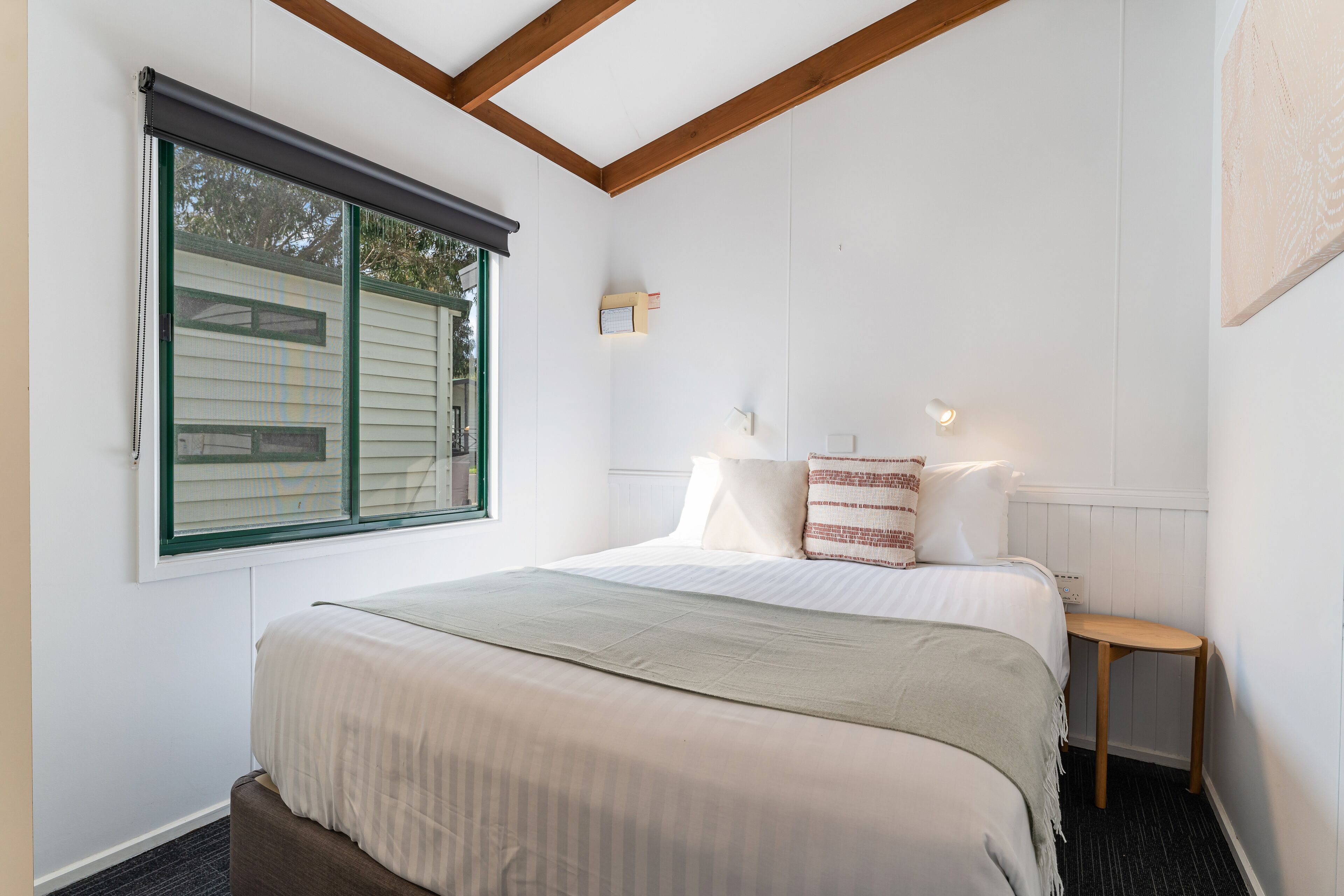 A neatly arranged bedroom with a large bed, a small bedside table, a window with blinds, and a wall-mounted light fixture.