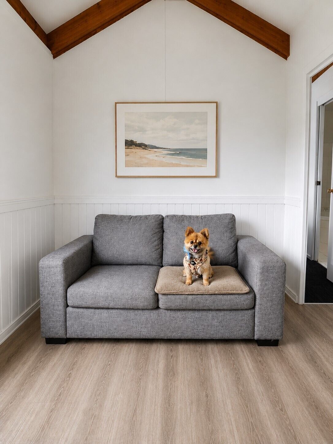 A grey sofa with a cushion, a framed picture of a beach, and a wooden floor.