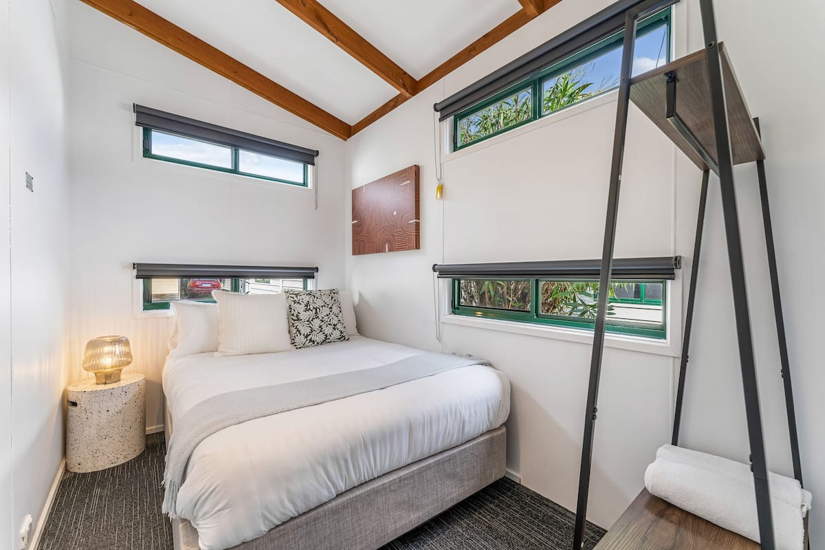 A bedroom with a bed, a bedside table with a lamp, a window with a view of greenery, and a ceiling with wooden beams.
