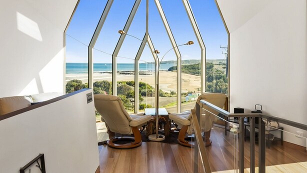 A room with a large glass window overlooking a beach and ocean. There are two chairs and a small table.