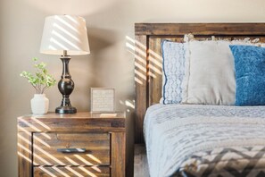 A neatly made bed with patterned bedding, a bedside lamp, a small plant, and a framed picture on a wooden nightstand. - Hot Tub, Outdoor pool, Pool & Air hockey (Hollister)